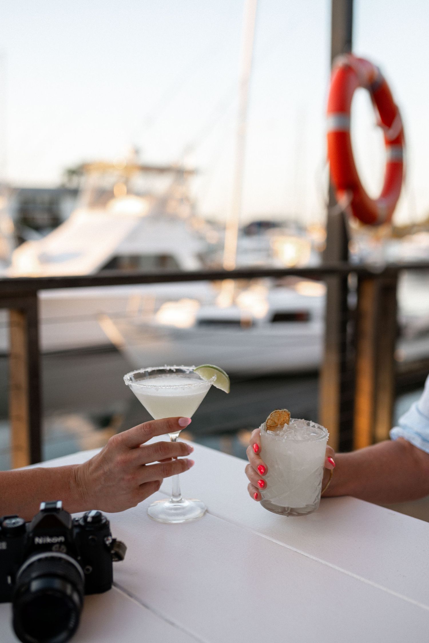 Two hands clinking cocktails at a marina dock bar with yachts and a life preserver ring in the background.