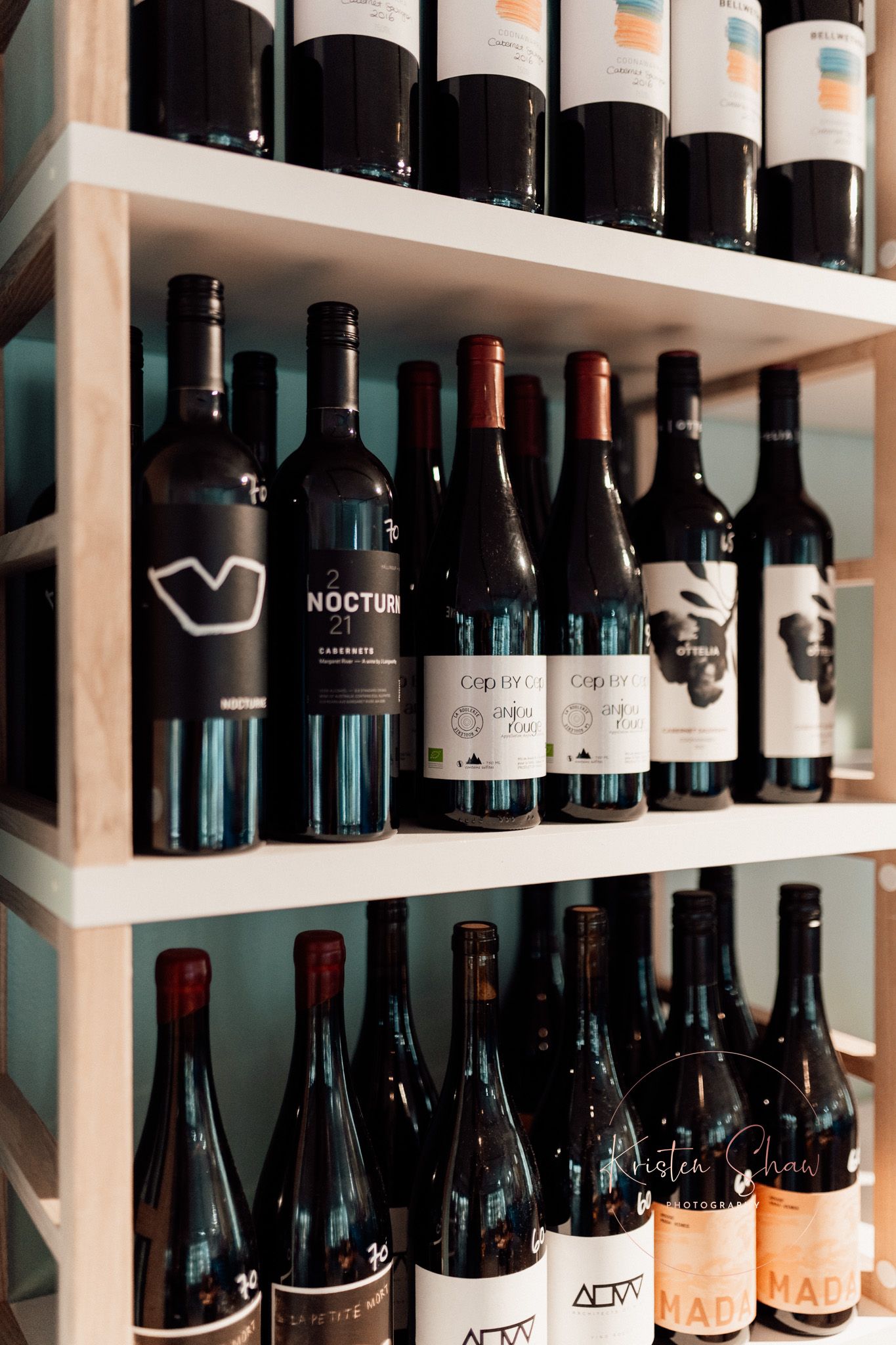 Wooden wine rack shelves displaying various red wine bottles including Nocturn Cabernet and Cop de Ra varieties in a wine shop.