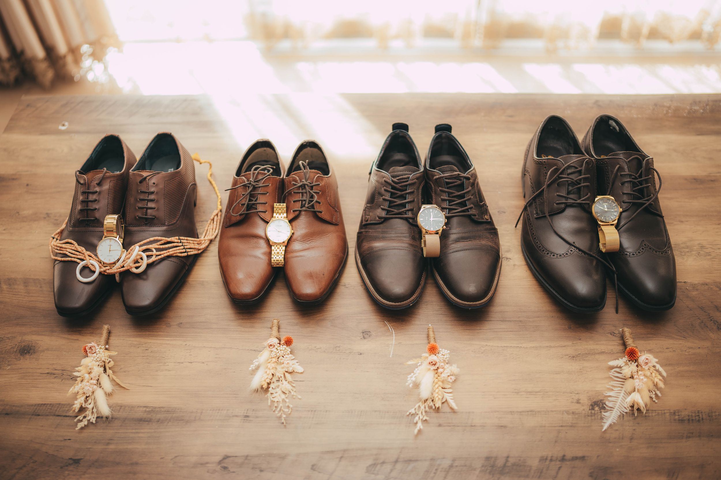 Four pairs of brown leather dress shoes with watches and dried flowers arranged on a wooden floor.