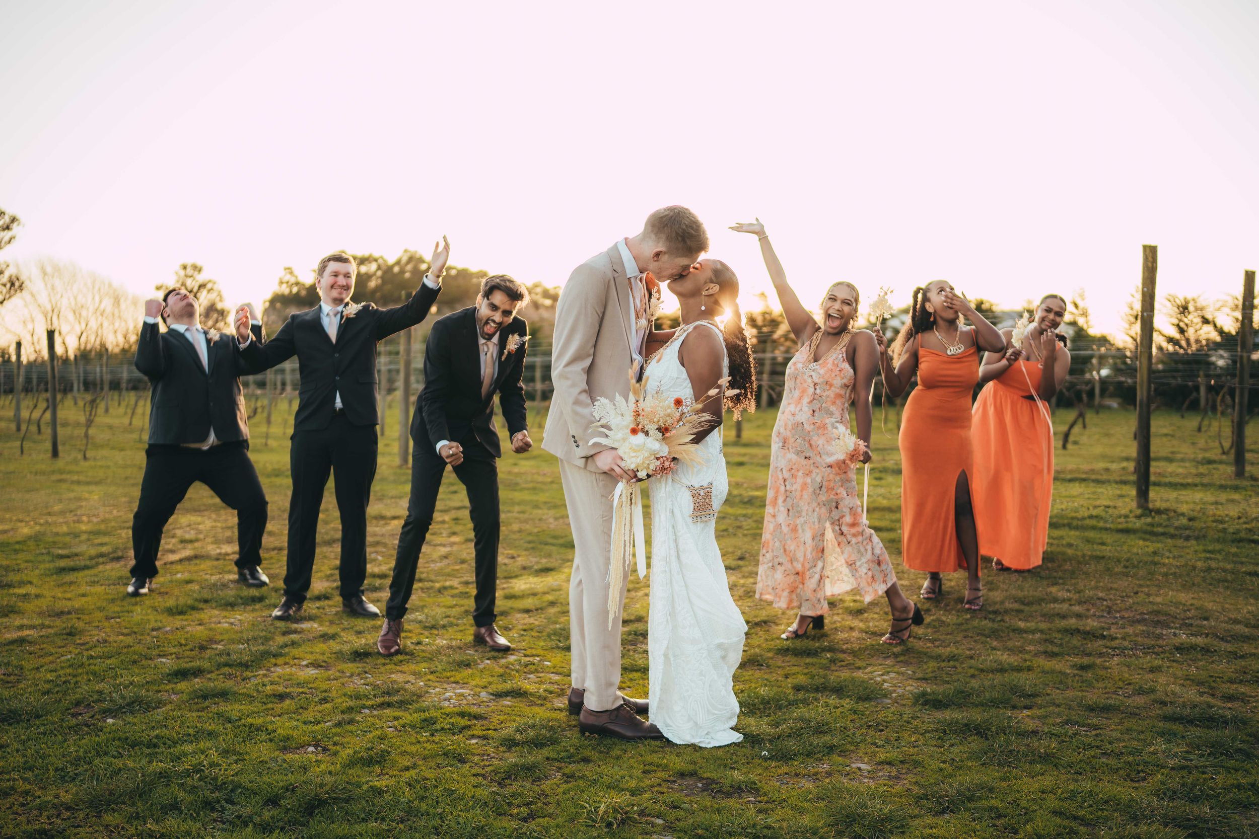 Bride and groom kissing at sunset vineyard wedding surrounded by cheering bridal party in colorful formal attire.