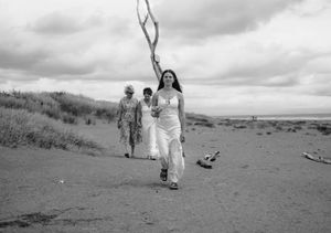 Black and white photo of women walking on a sandy beach path with driftwood and cloudy skies.