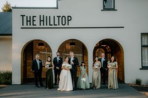 Wedding party poses outside The Hilltop venue, bride and groom centered with groomsmen and bridesmaids in sage green gowns.
