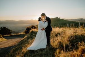 Bride and groom share a romantic kiss at golden sunset on a scenic hilltop with rolling mountains in the background.