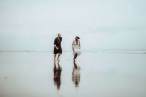 Two women walking barefoot on a reflective wet beach shore on an overcast day, wearing black and white dresses.
