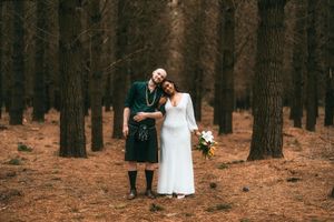 Couple in wedding attire poses in pine forest, groom in green kilt, bride in white gown holding bouquet.