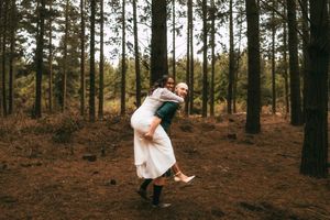 Couple sharing a joyful piggyback ride in a pine forest, wearing wedding attire, surrounded by tall trees.