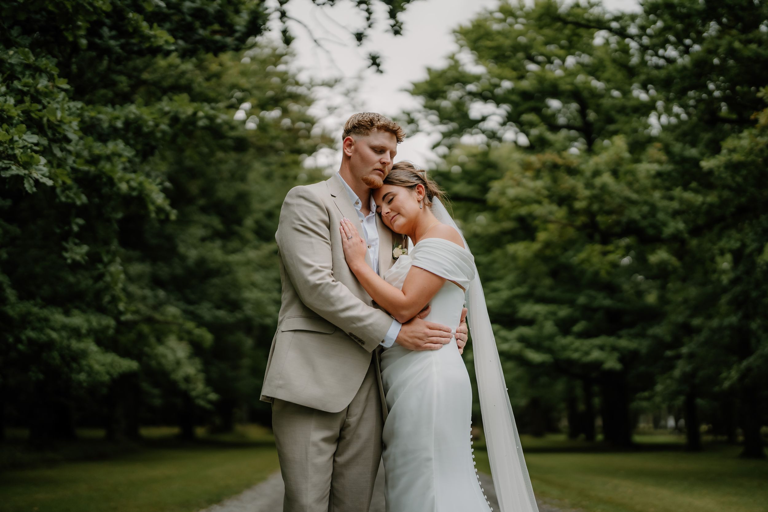 A bride and groom share a romantic embrace on a tree-lined path, wearing a white gown and tan suit.