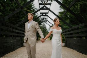 Bride and groom hold hands on a rustic bridge, she in a white off-shoulder gown, he in a tan suit.