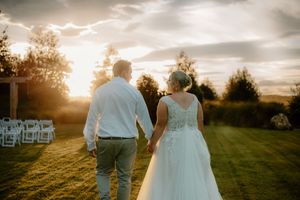 Bride and groom holding hands walking toward sunset at outdoor wedding venue with white chairs.