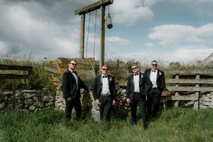 Four groomsmen in black tuxedos and bow ties pose confidently near rustic stone wall and wooden gate.