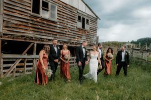 Wedding party walks beside rustic barn, bride in white gown with bridesmaids in terracotta dresses and groomsmen in black tuxedos.
