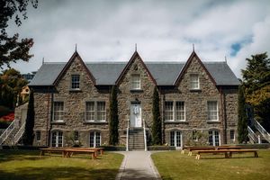 Historic stone manor house with Gothic architecture, manicured lawn, and wooden benches under cloudy skies.