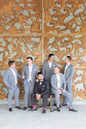 Groom in black tuxedo seated among groomsmen in gray suits with bow ties, posing in front of ornate copper wall art.