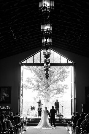 Black and white wedding ceremony photo of bride and groom at altar before large window with tree backdrop.