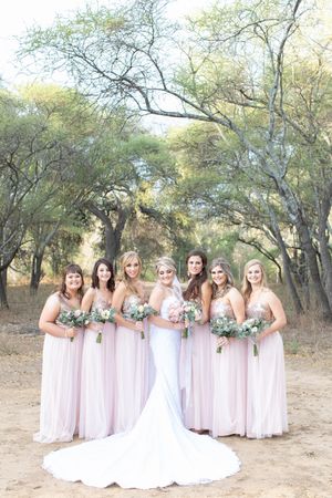 Bride in white gown with six bridesmaids in blush pink dresses holding bouquets outdoors among trees.