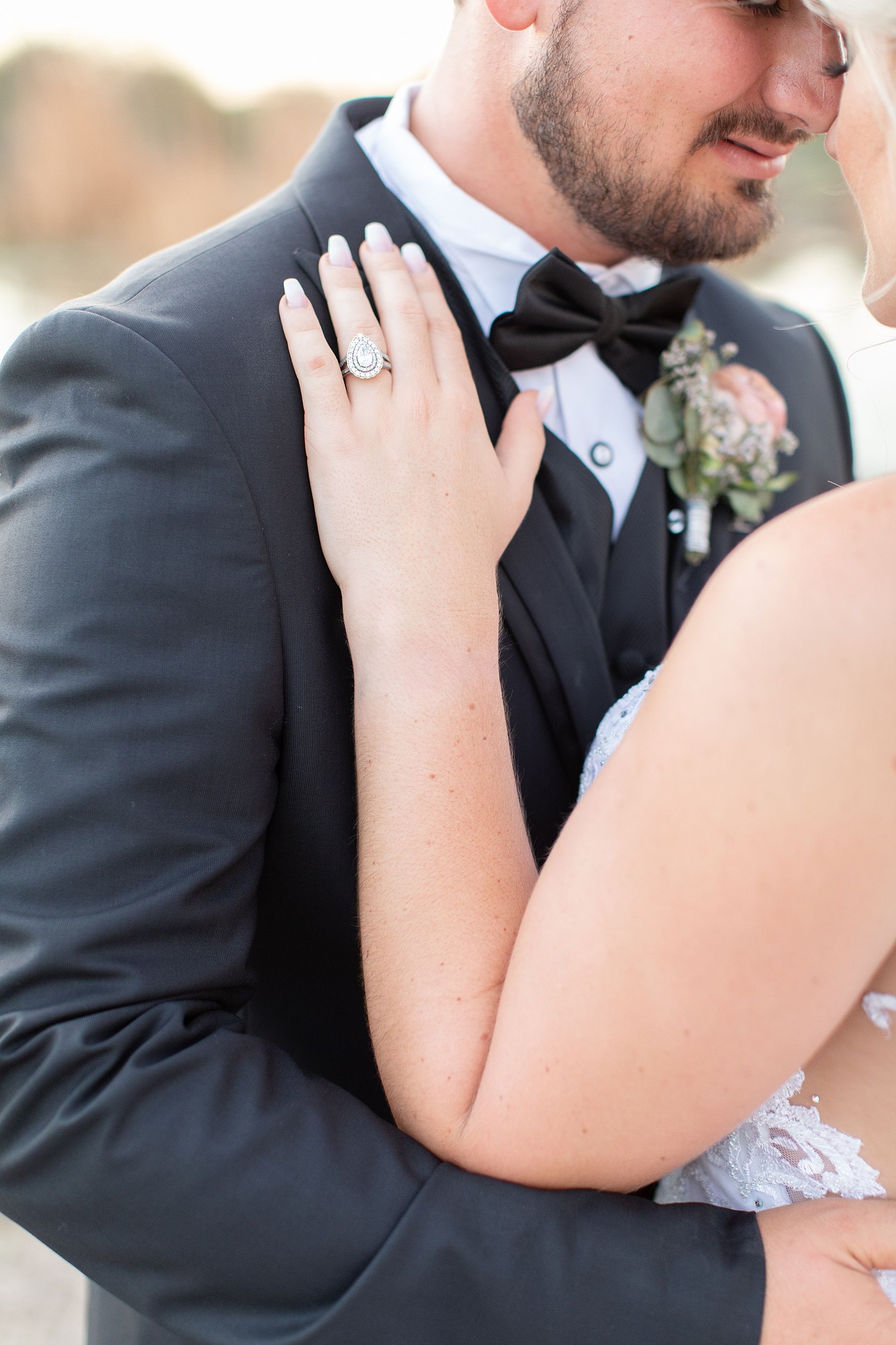 Bride resting hand on groom's tuxedo lapel, showcasing elegant engagement ring, with floral boutonniere detail.