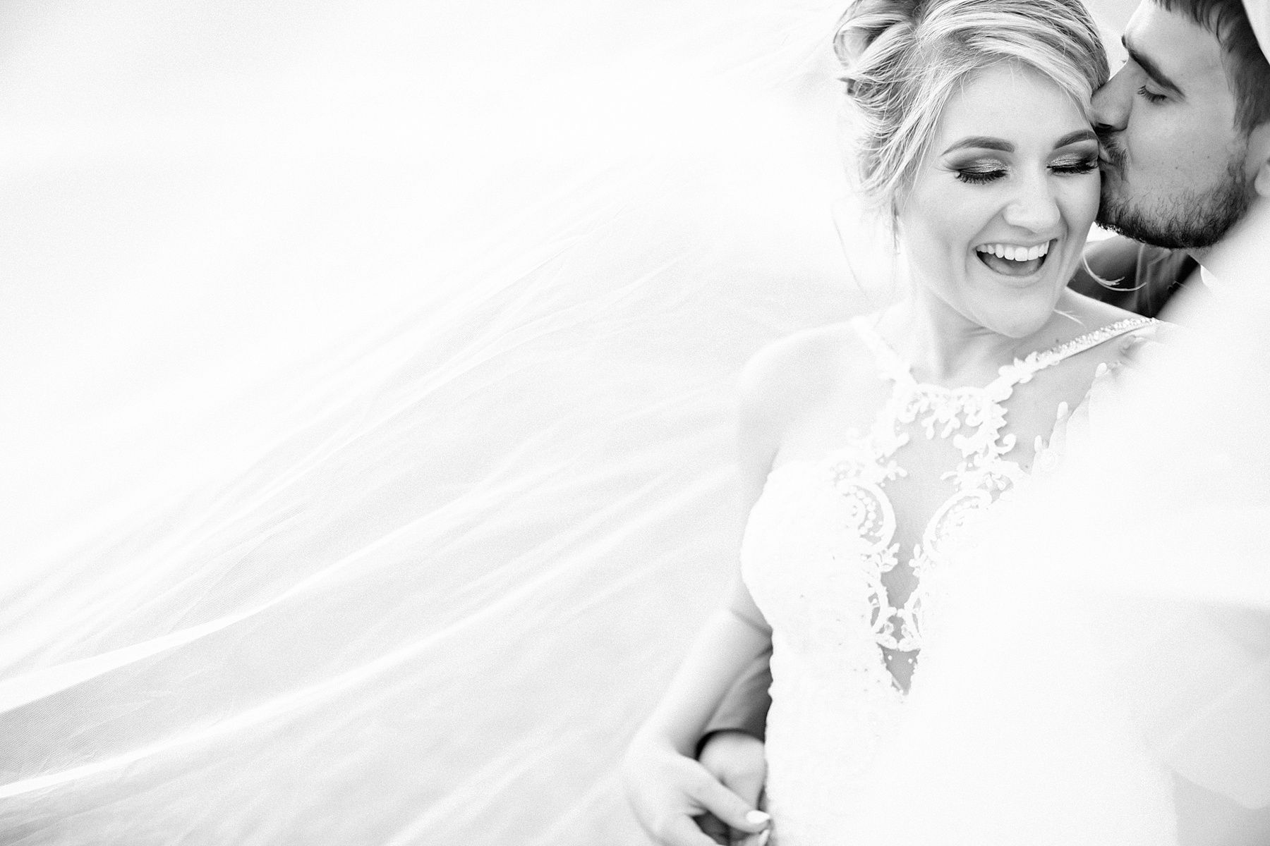 Black and white wedding photo of a laughing bride in lace gown with flowing veil as groom kisses her cheek.