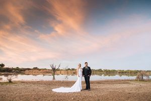 Bride and groom stand by a serene lake at sunset with dramatic pink clouds in the sky on their wedding day.