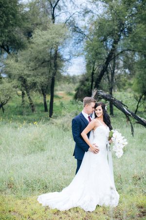 Bride and groom embrace in a lush green meadow, bride wearing a white gown holding a bouquet, groom in a navy suit.