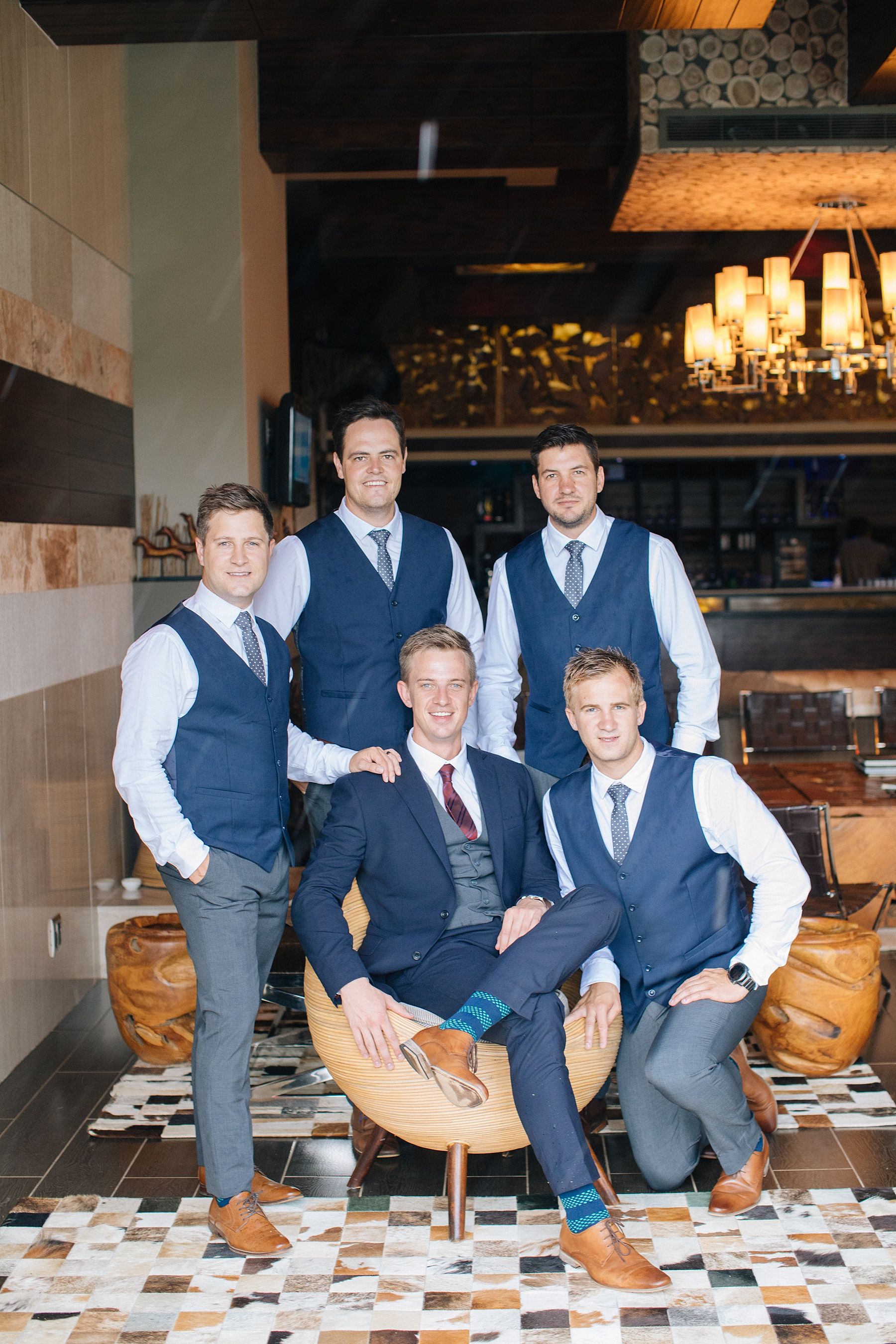 Five groomsmen in navy vests and suits pose together in an elegant restaurant setting for a wedding photo.