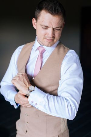 Groom in beige vest and pink tie adjusting cufflinks on white dress shirt while wearing a gold watch.