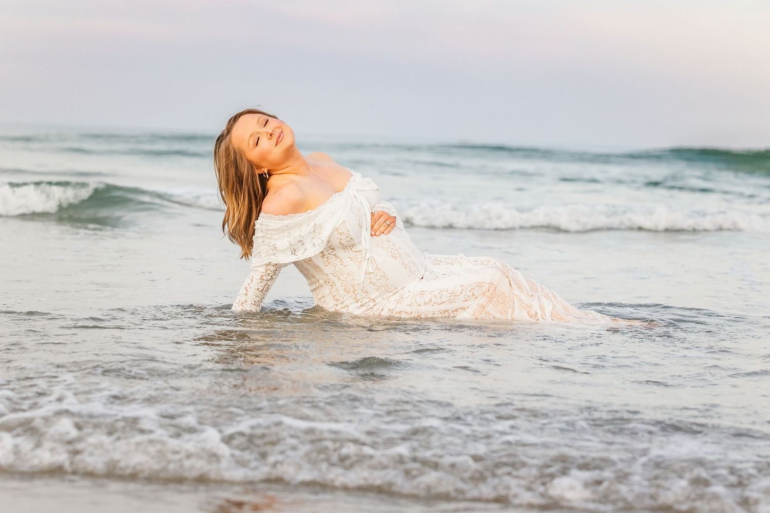 Pregnant woman in white lace dress posing gracefully in ocean waves on a serene beach at sunset.