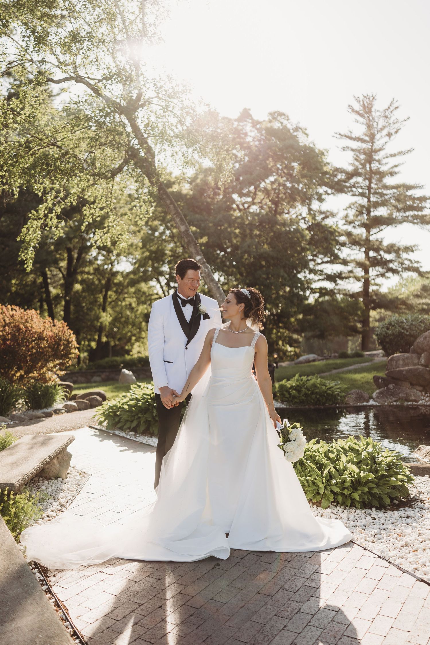 Bride and groom hold hands outdoors in sunlit garden, she in white gown, he in white tuxedo jacket.