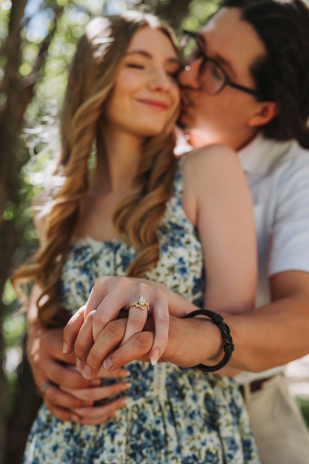 Couple in a sunny park, woman in floral dress showing engagement ring while man kisses her cheek.