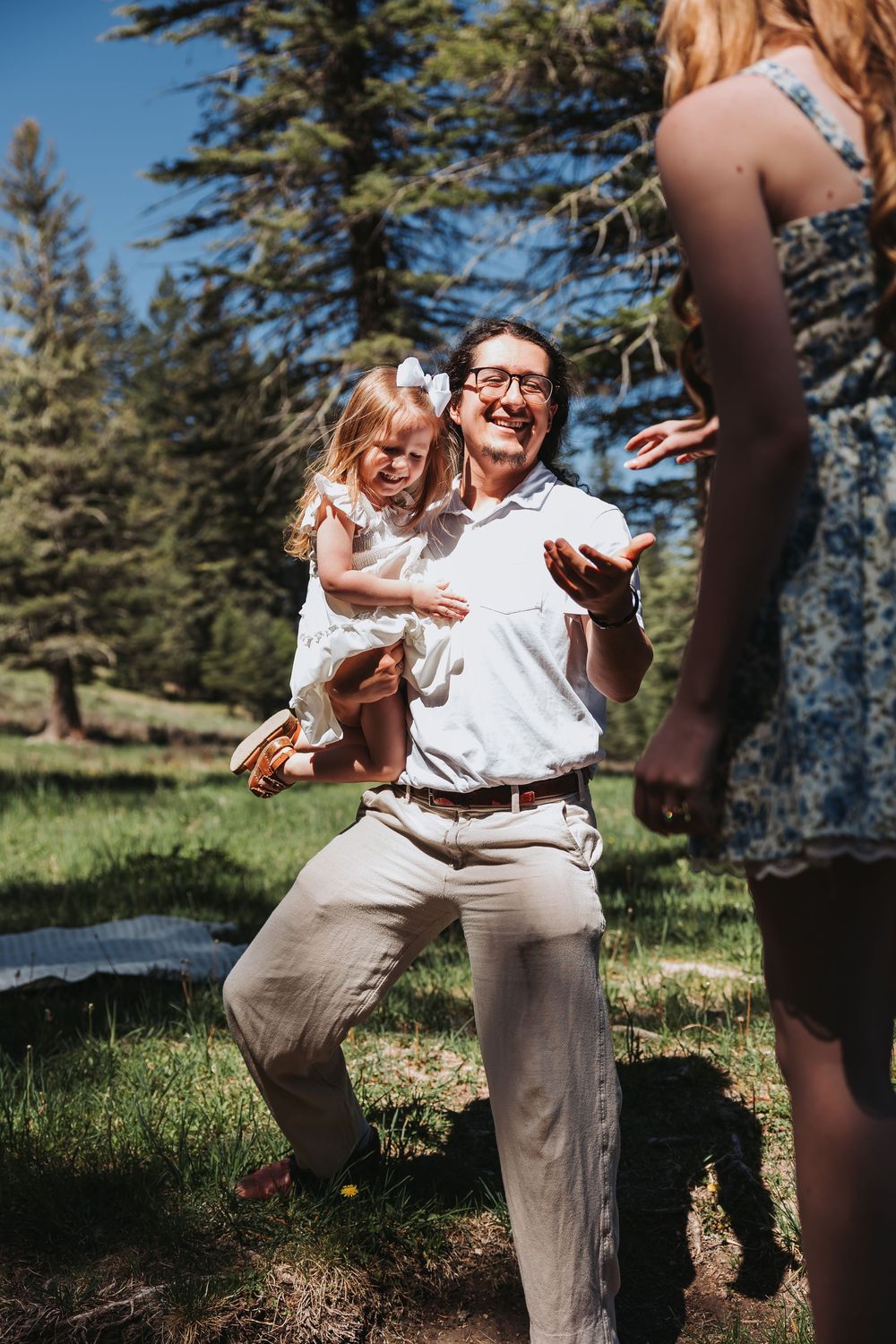 A laughing man holds a little girl in a sunny forest clearing while reaching toward a woman in a floral dress.