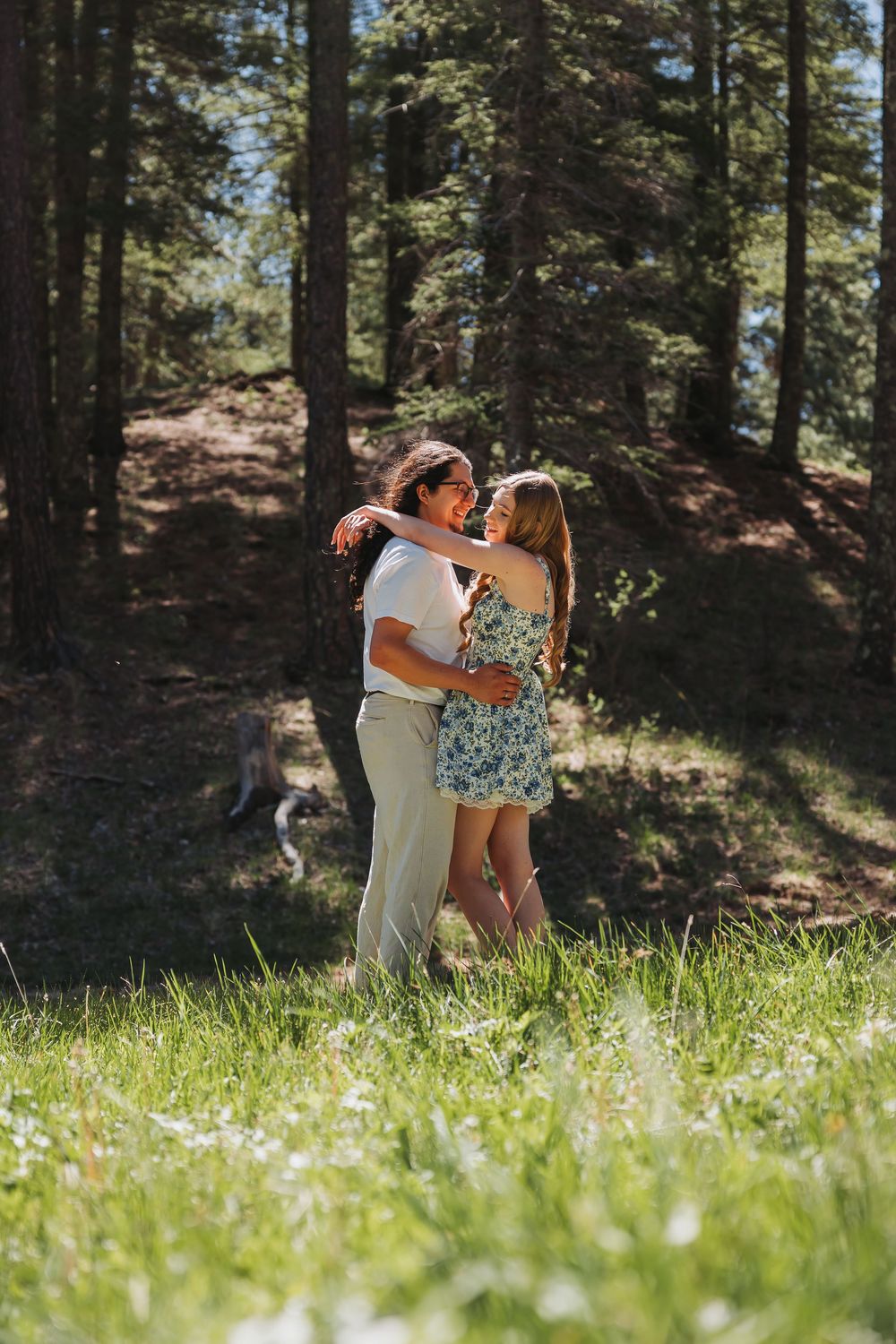 A couple embraces and smiles at each other while standing in a sunlit forest meadow with tall pine trees.