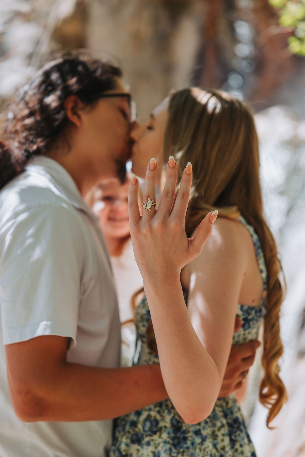 Couple kissing during engagement moment while woman displays stunning diamond ring on her hand outdoors.
