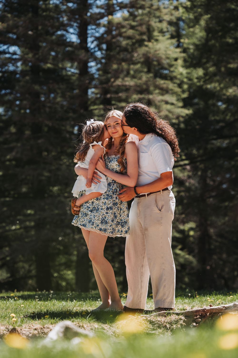 Two women and a child sharing a tender moment outdoors in a sunny forest meadow.