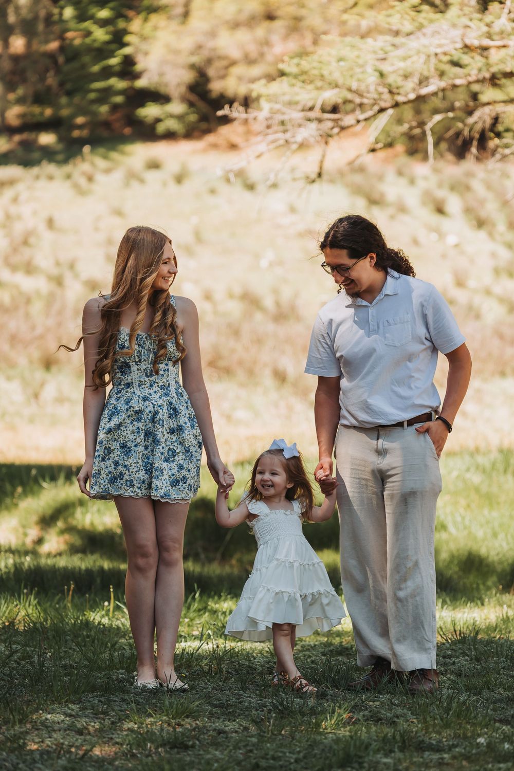 A young family with a toddler girl in white dress holds hands while walking on green grass outdoors.