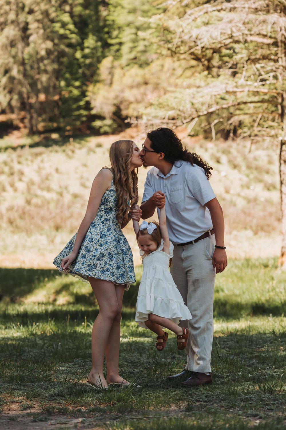 A couple kisses while swinging their toddler daughter between them in a sunny green park.