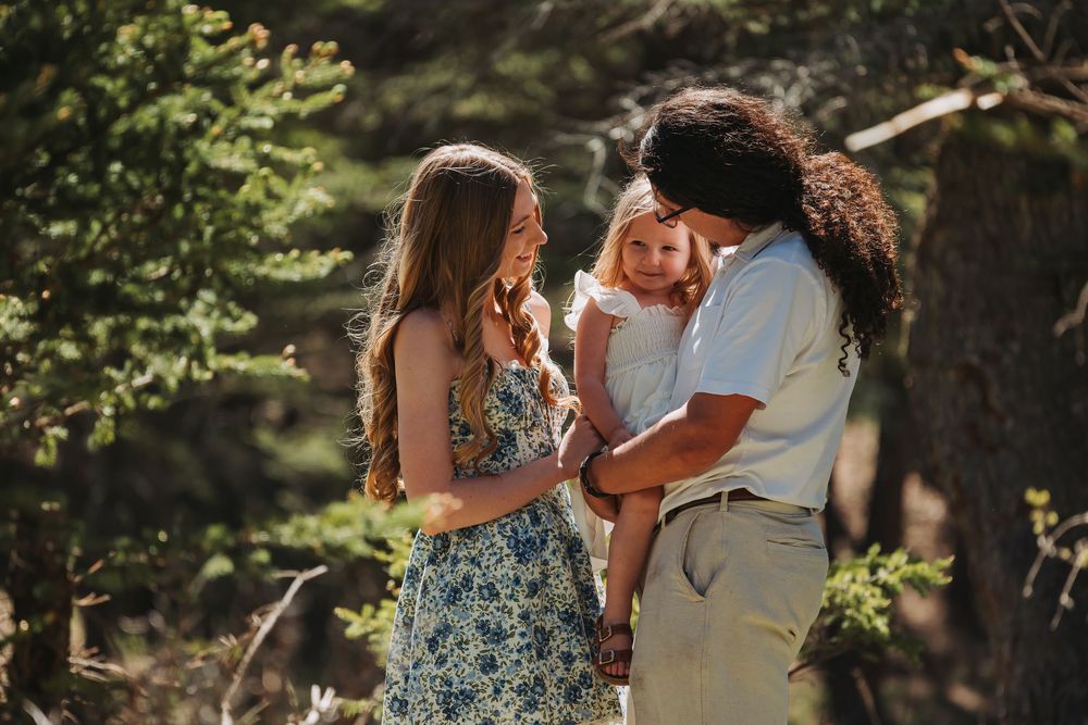 A young family with a baby enjoys an outdoor moment among pine trees in a sunlit forest setting.