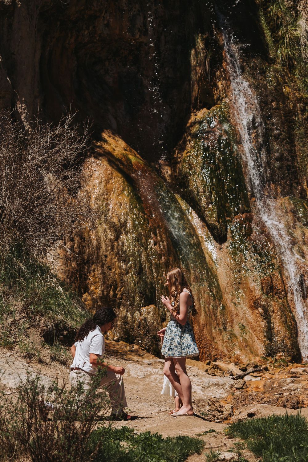 Man proposing to woman on one knee near a scenic waterfall in a natural rocky outdoor setting on a sunny day.