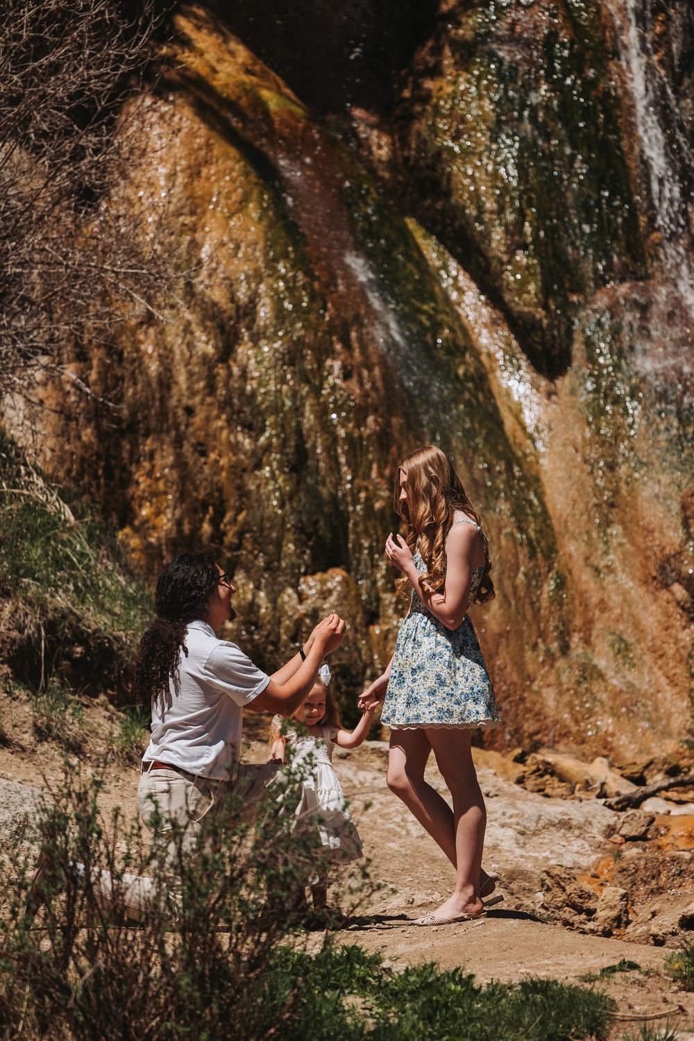 Man proposing to woman on one knee near a scenic waterfall on rocky terrain outdoors.