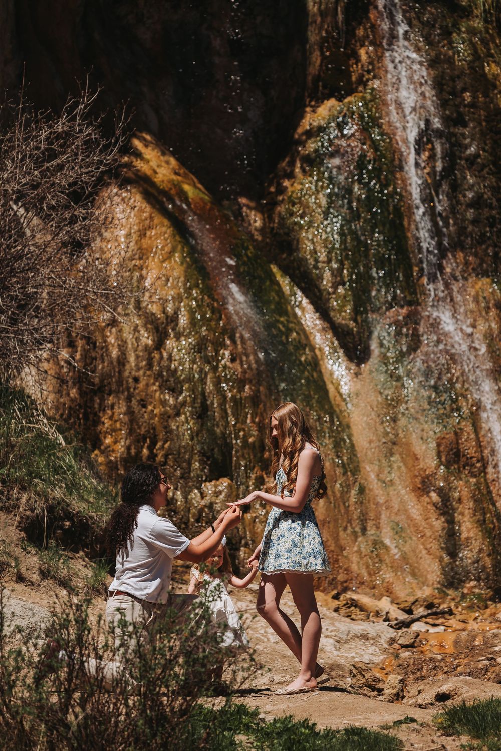 Man proposing to woman in floral dress by scenic waterfall on rocky terrain in bright sunlight.