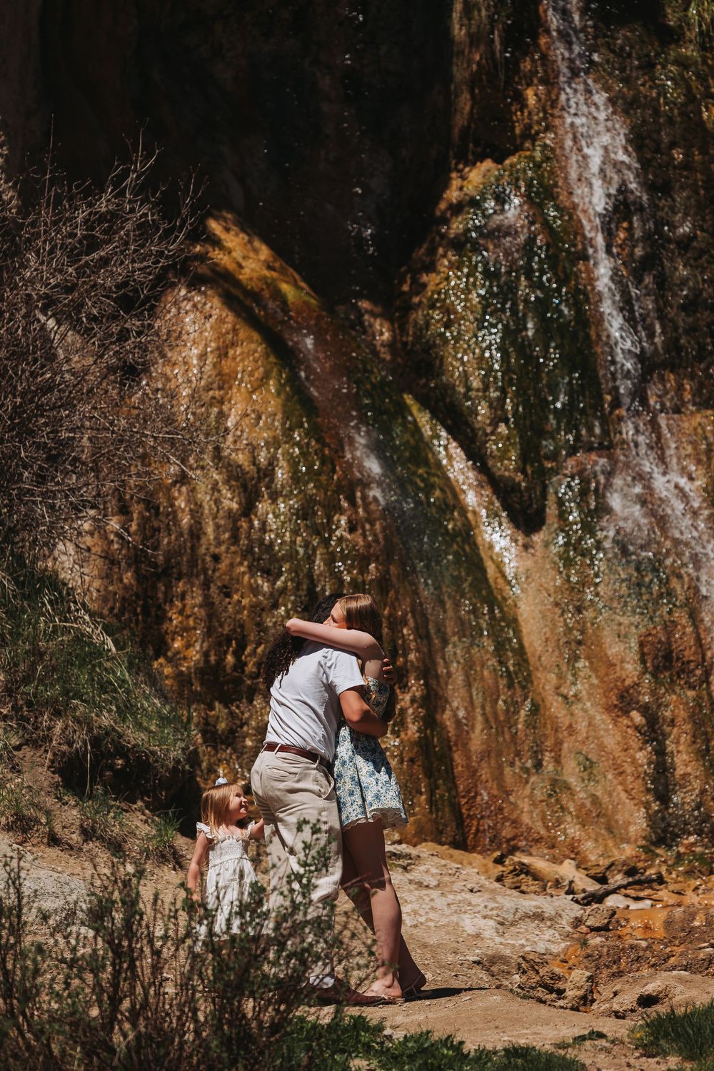 Family enjoying a scenic waterfall hike, with a parent lifting a child near a mossy rock face outdoors.