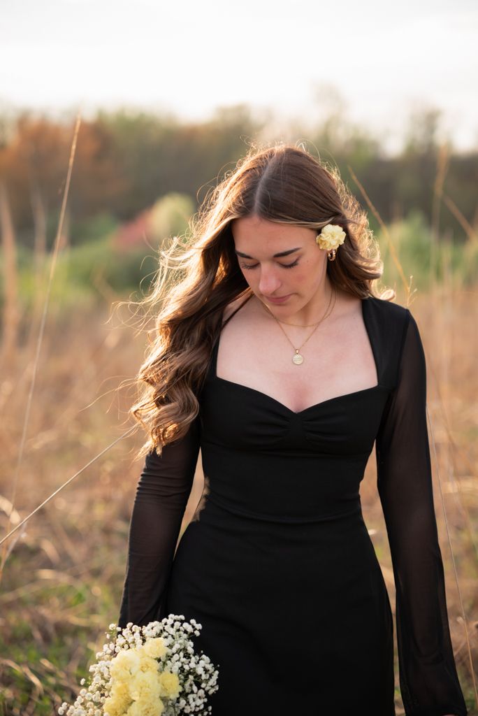 Young woman in black dress holding white flowers in golden field at sunset, with flower in hair.