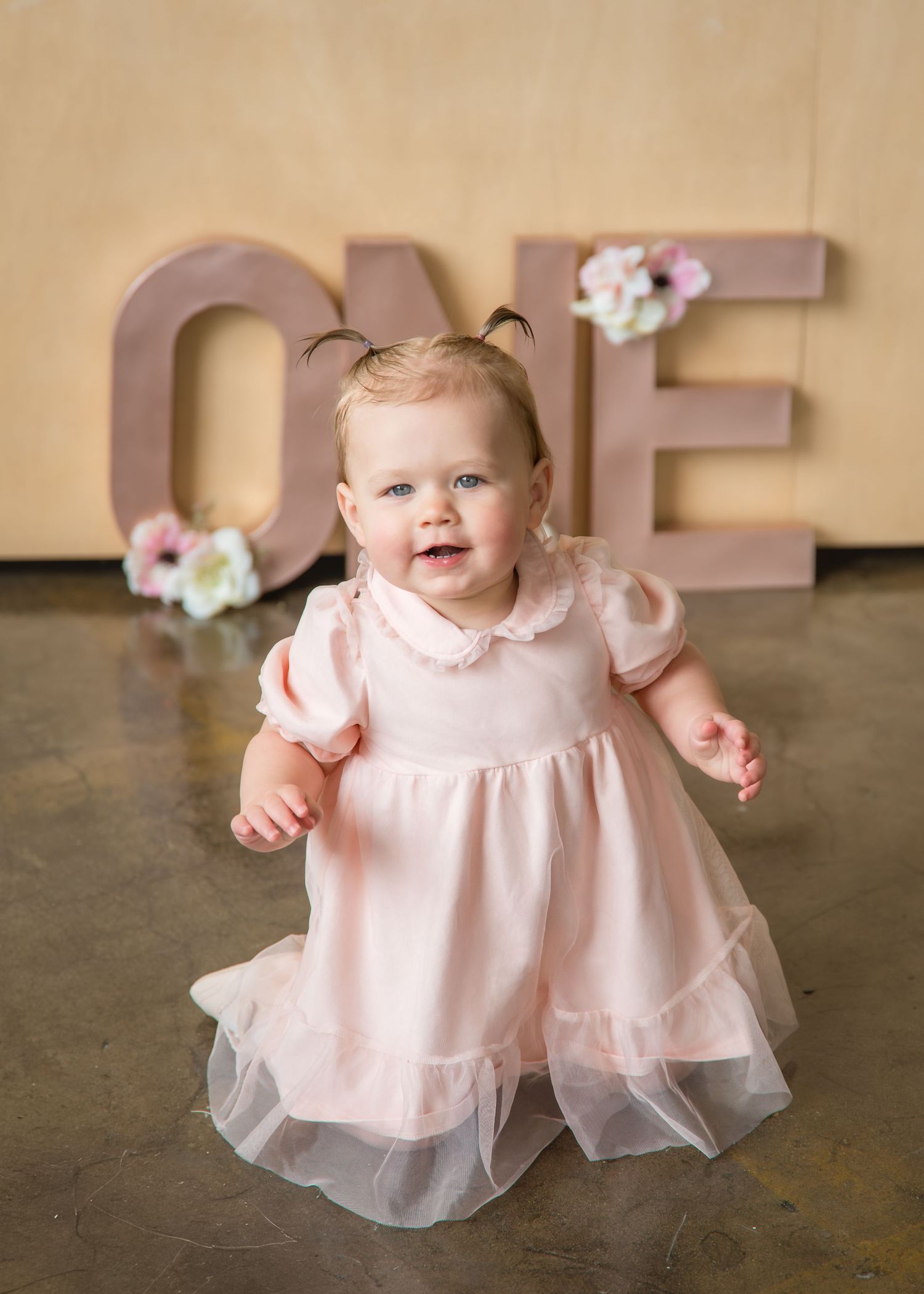 A smiling baby girl in a pink tulle dress stands in front of large ONE letters for her first birthday photo.