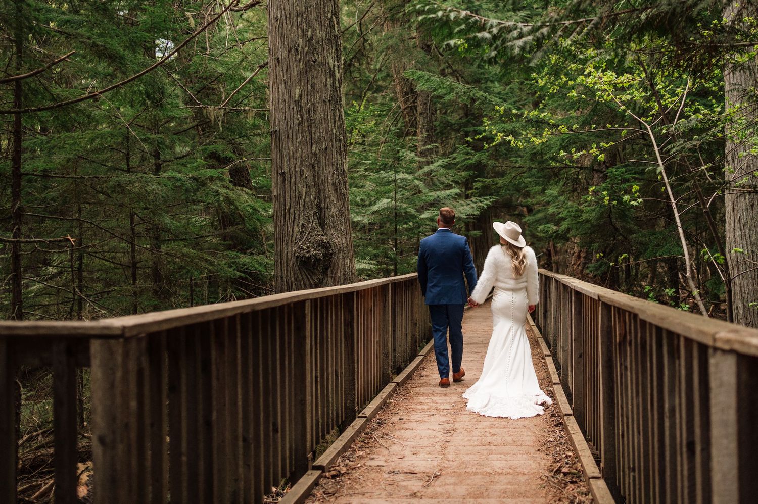 Newlywed couple walking hand-in-hand on a wooden bridge surrounded by lush green forest trees.