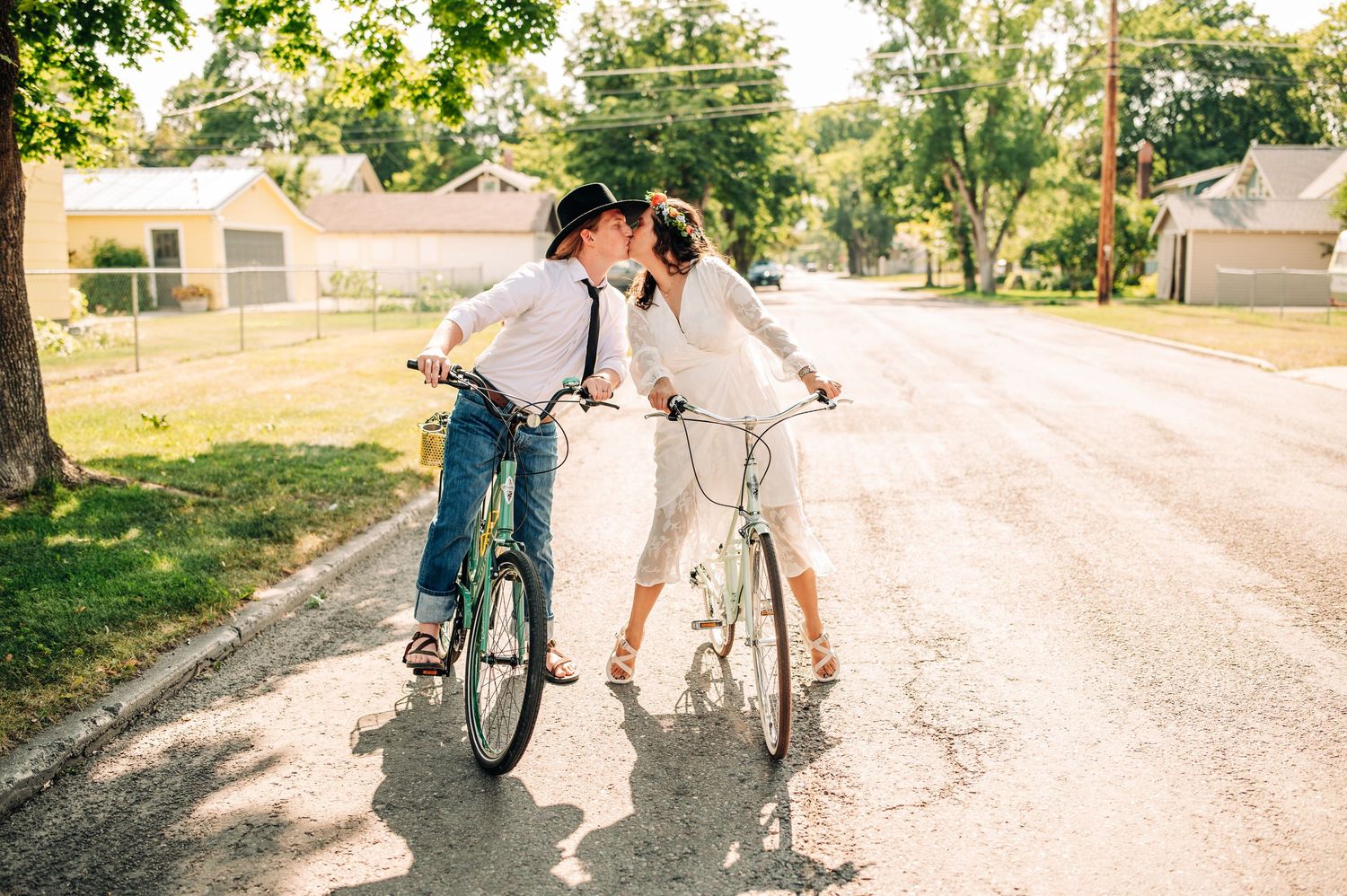 A couple sharing a romantic kiss while riding bicycles on a sunny suburban street lined with trees.