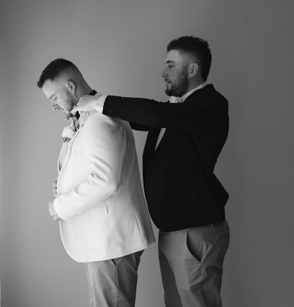 Black and white photo of two men in suits, one adjusting the other's jacket collar in a formal wedding setting.