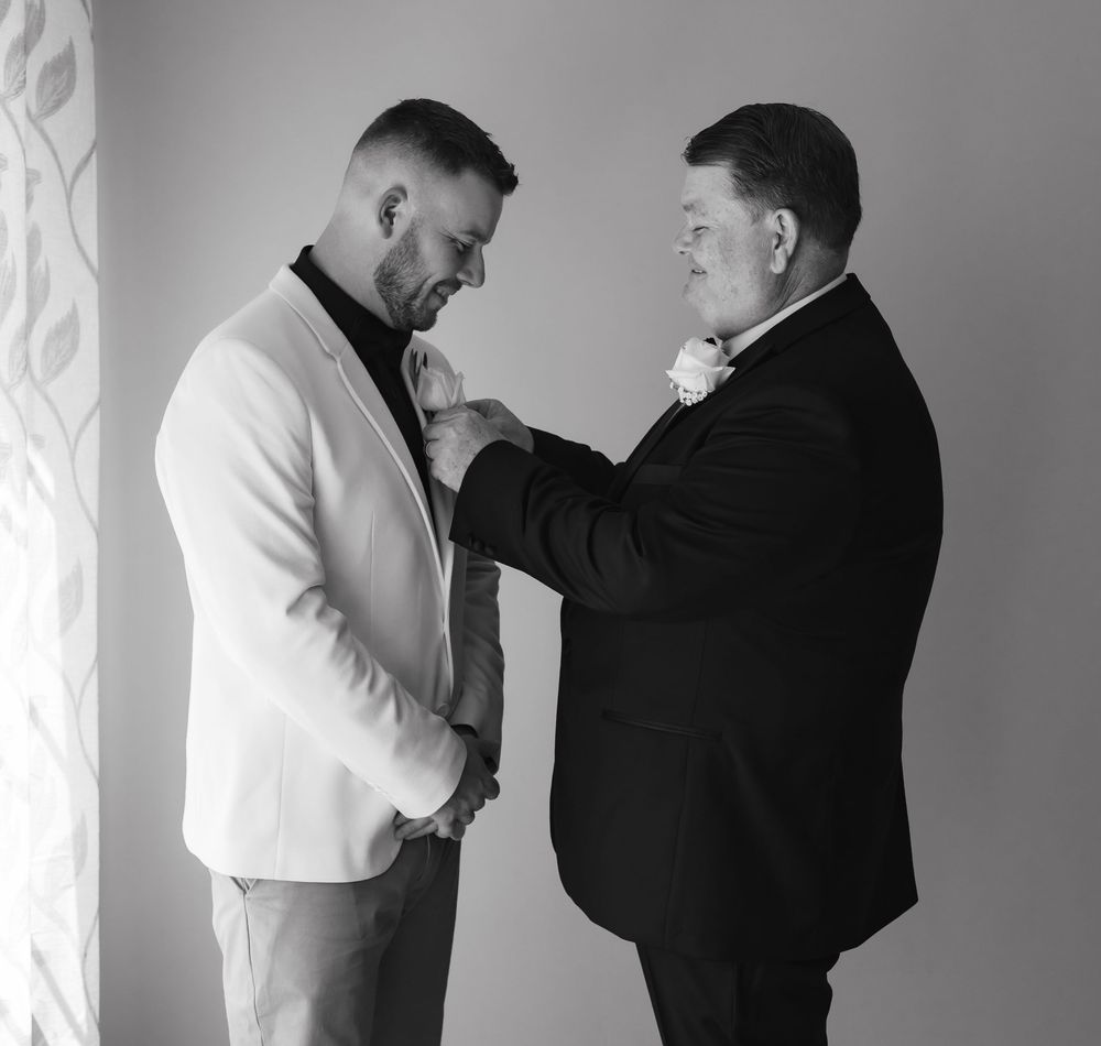 Black and white photo of two men in suits, one adjusting the other's boutonniere before a wedding ceremony.