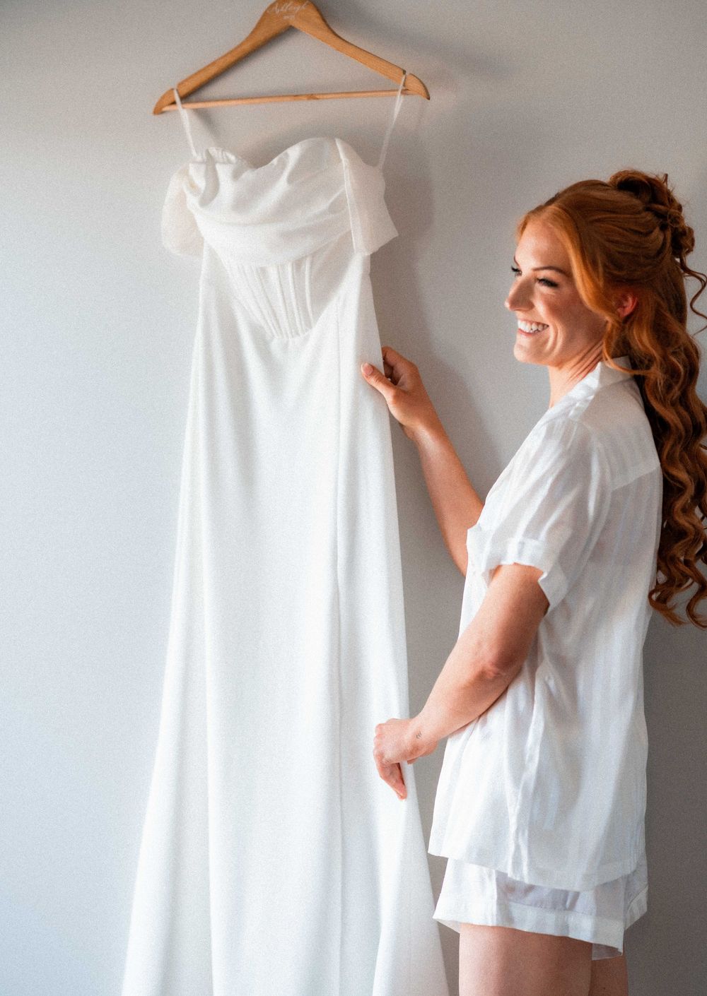 Smiling redhead bride in white robe admiring her elegant off-shoulder corset wedding gown on hanger.