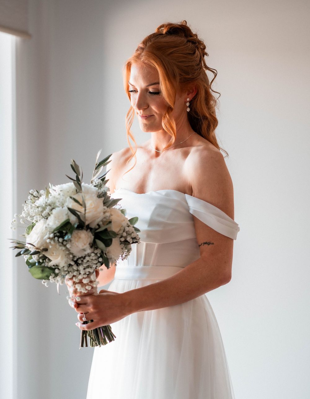 Elegant bride in off-shoulder white gown holding white floral bouquet, with red hair and pearl earrings.