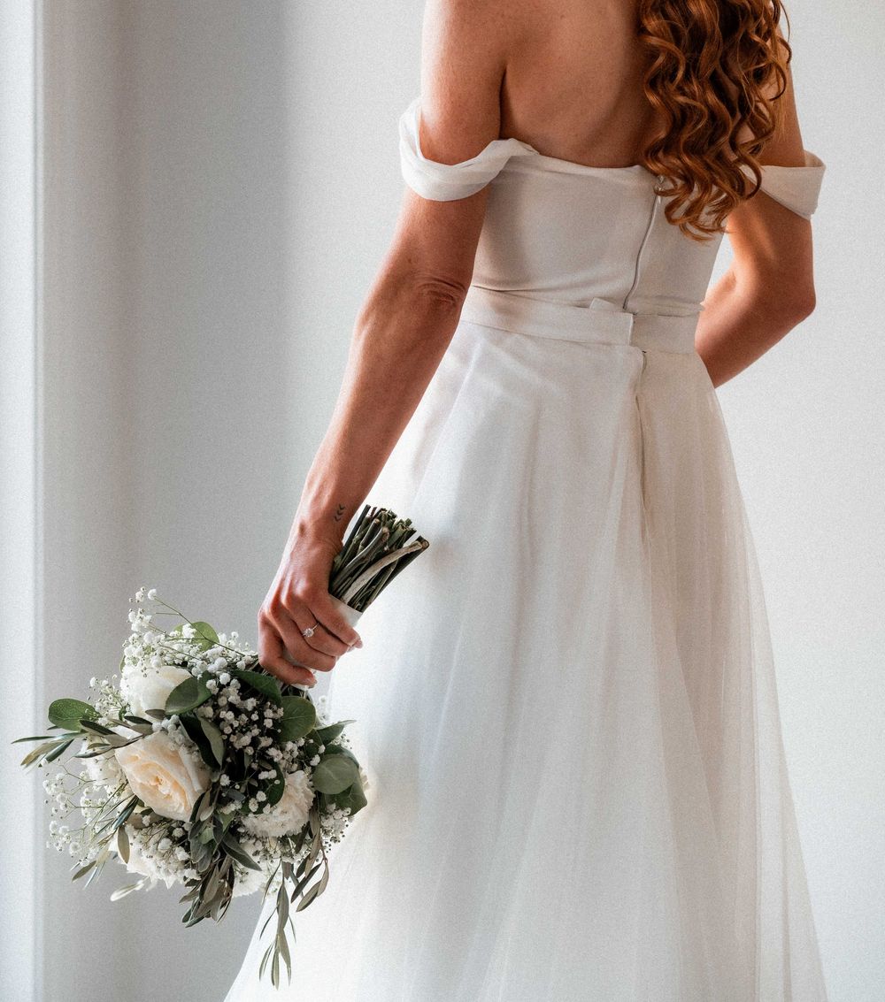 Bride in off-shoulder white tulle wedding gown holding elegant bouquet of white roses and greenery, back view.