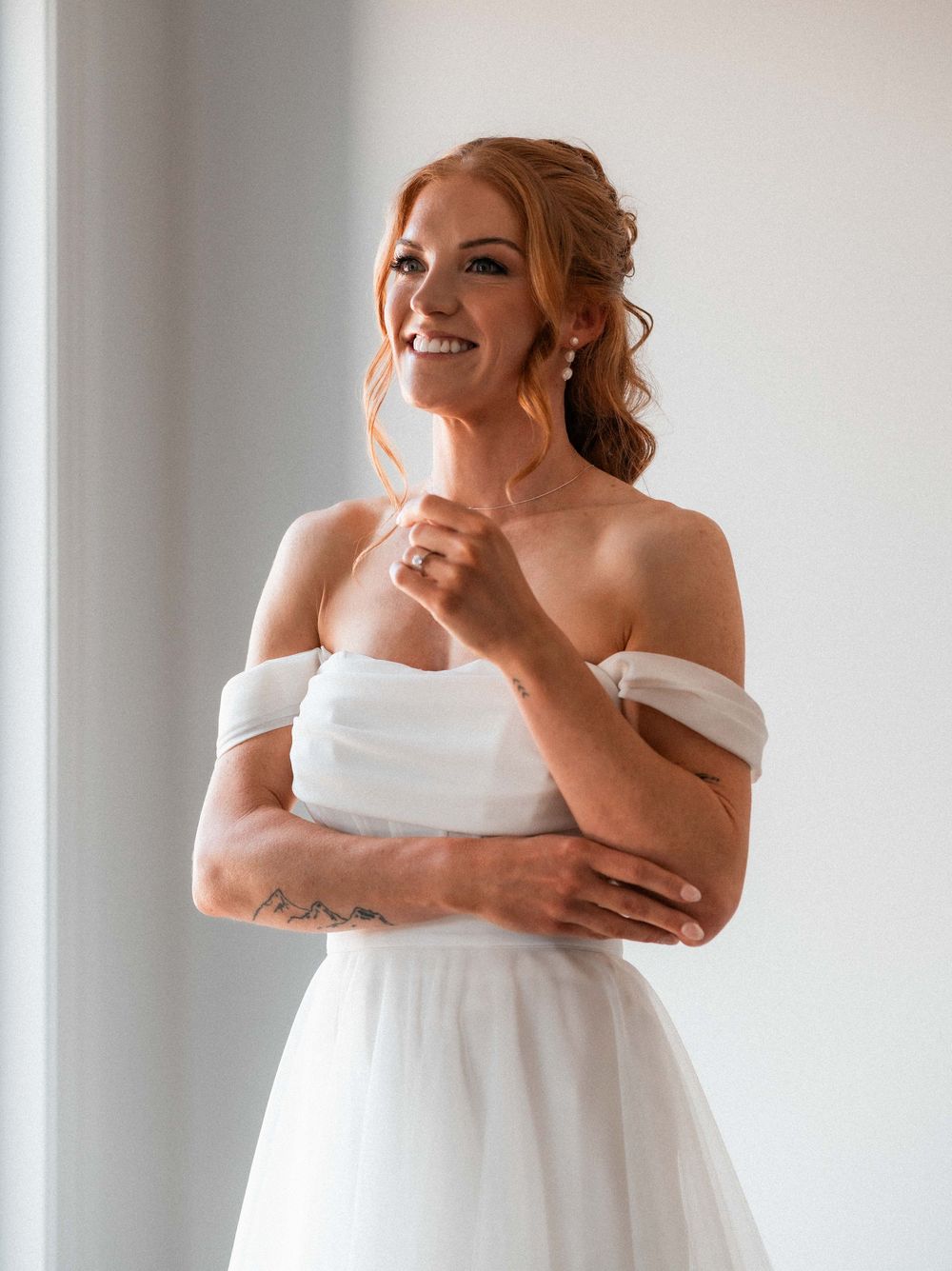 Smiling bride in off-shoulder white gown with red hair up, wearing earrings and ring, arms crossed near bright window.