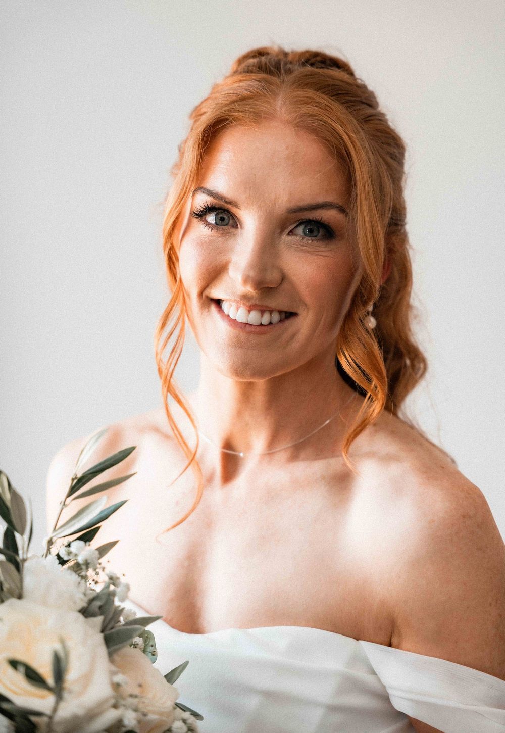Smiling bride with red hair holding white flowers, wearing an off-shoulder gown and pearl earrings.